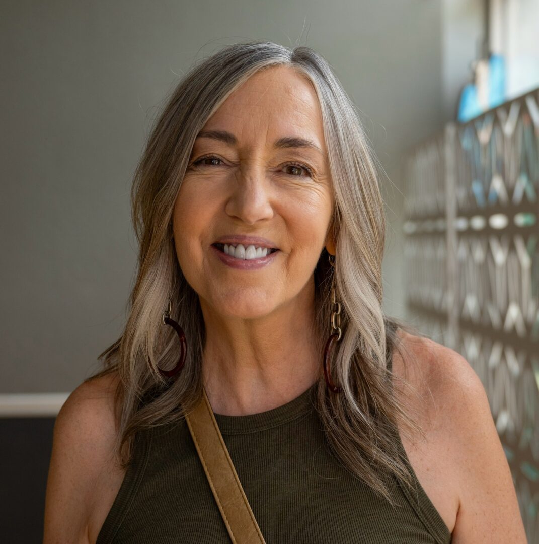 Smiling woman with long gray hair and a tank top stands indoors near a patterned wall.