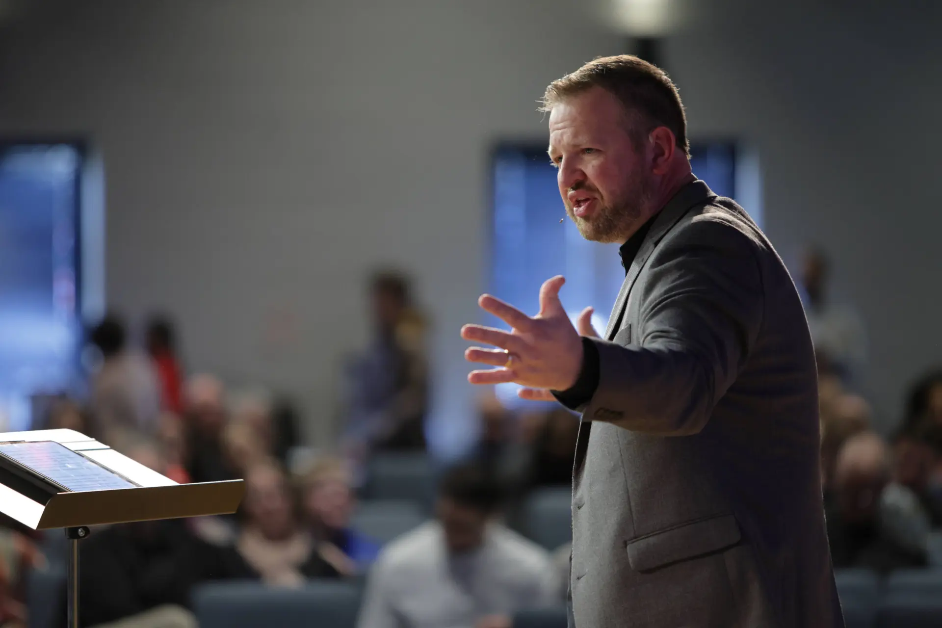 A man in a suit gestures while speaking at a lectern in front of an audience in a blurred indoor setting.