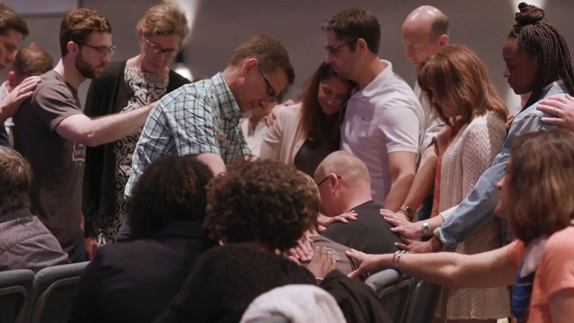 A group of people gather closely, placing their hands on one person seated in the middle, appearing to offer support or comfort in a communal setting.