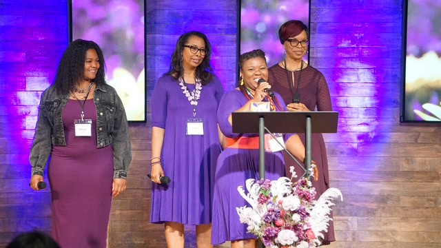 Four women in purple dresses stand on a stage with a microphone and floral arrangement. Three wear glasses and lanyards. Background features purple-lit panels with floral imagery.