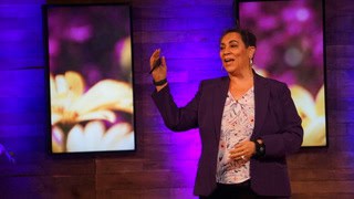 A person in a purple blazer speaks enthusiastically on stage, with two screens displaying blurred floral images in the background.