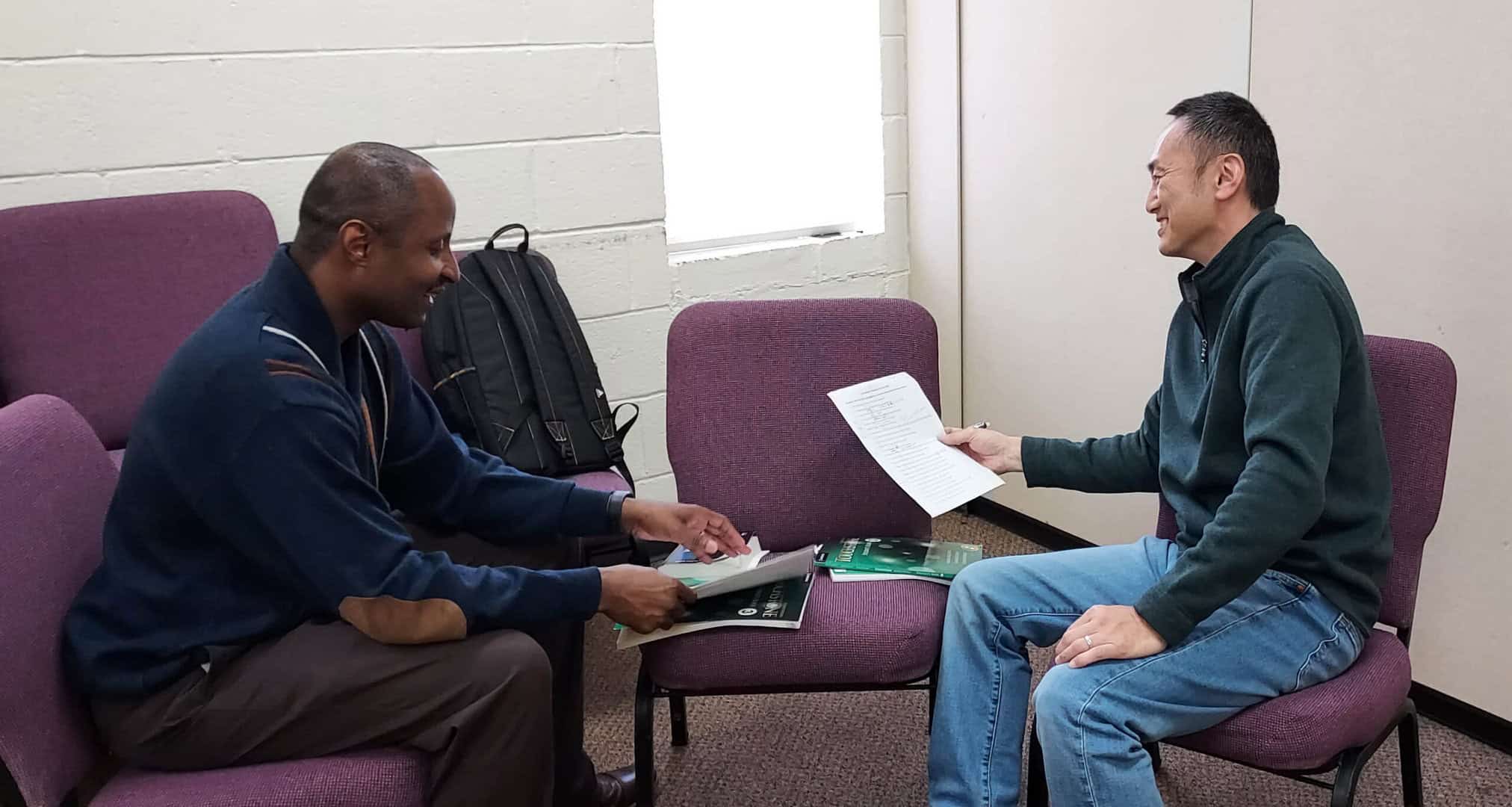 Two men seated in a room with purple chairs, engaged in conversation. One holds documents, while the other gestures with a paper. A backpack is on a chair nearby.