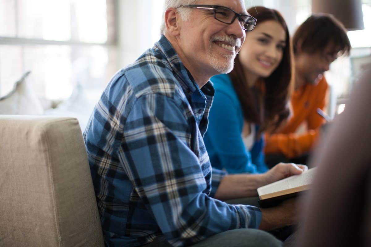 A smiling man in a plaid shirt sits on a couch holding a book, while a woman and another person are seated nearby.