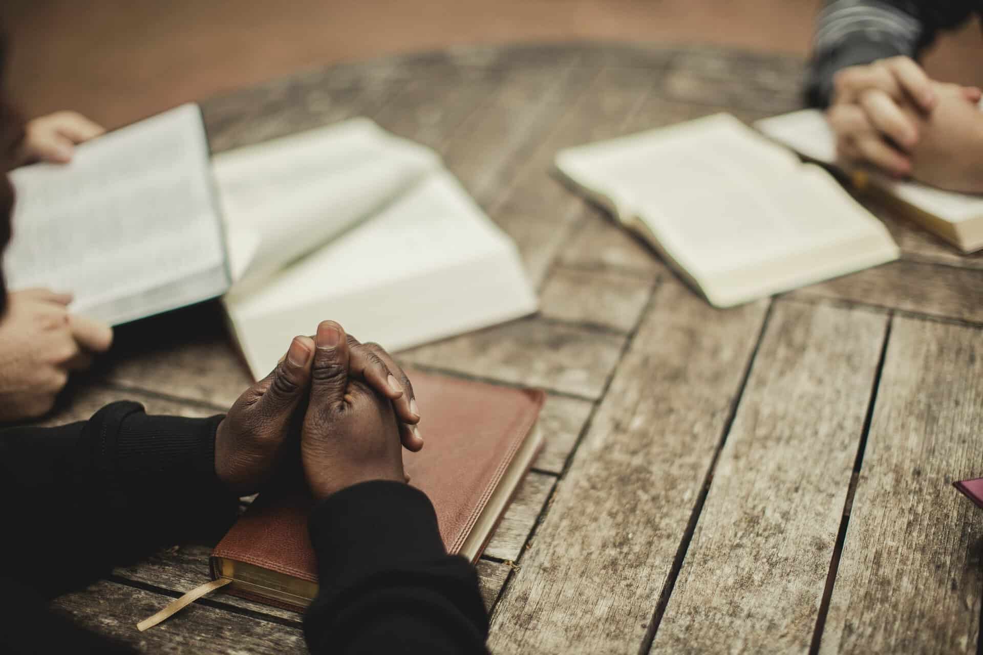 Three people sit around a wooden table with open books, two of them rest their hands on top of the books.