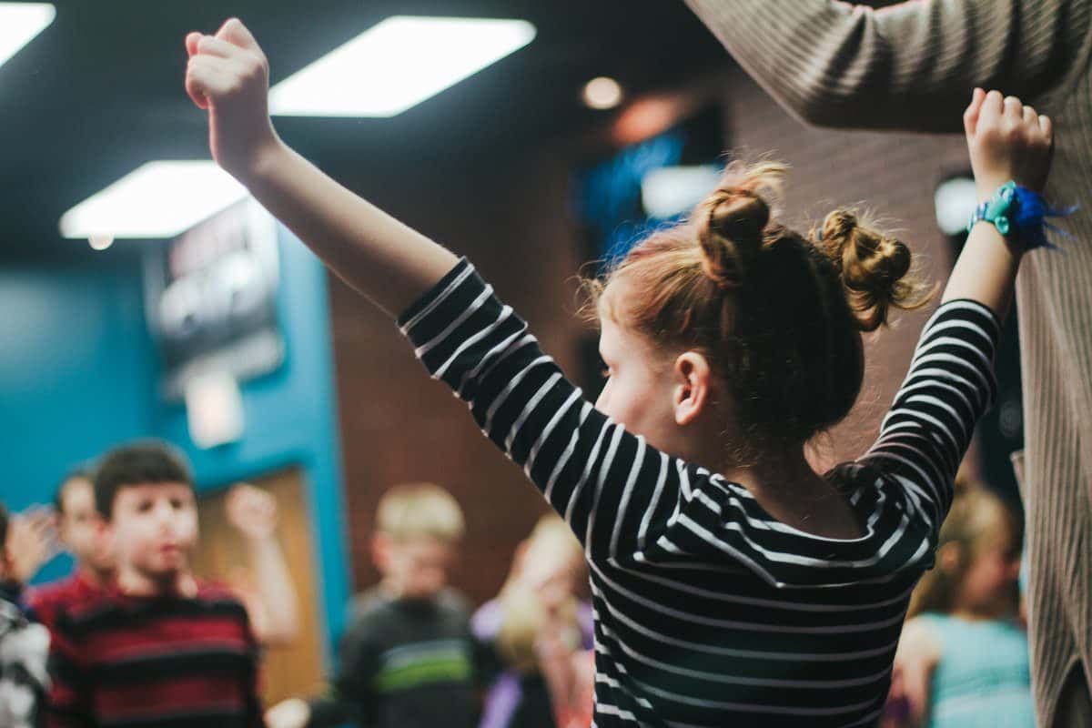 Child with raised arms participates in an indoor activity, surrounded by other children and adults.