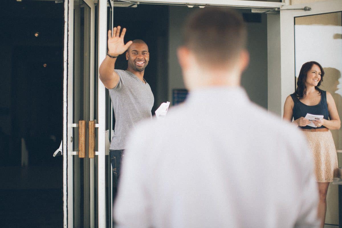 A man waves at a person entering the building, which feels like a second home, while another person stands nearby holding papers.