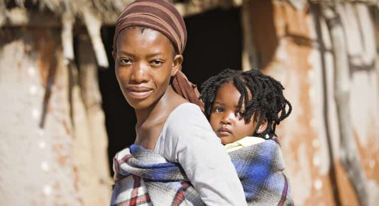 A woman stands outdoors, carrying a child on her back wrapped in a plaid cloth. The backdrop hints at a rustic building, evoking scenes from distant missions.