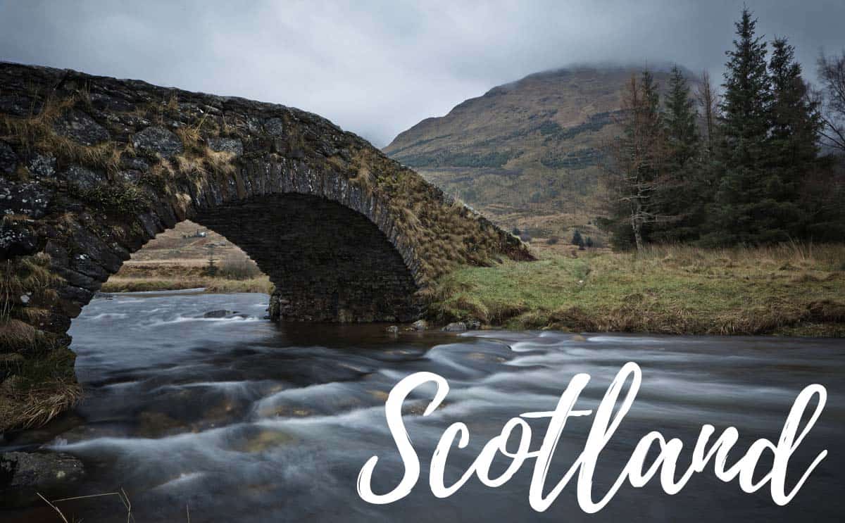 An ancient stone bridge arches over a flowing river in a mountainous, cloudy landscape, hinting at the distant missions of time. "Scotland" is elegantly scripted at the bottom, evoking an aura of exploration akin to space missions.